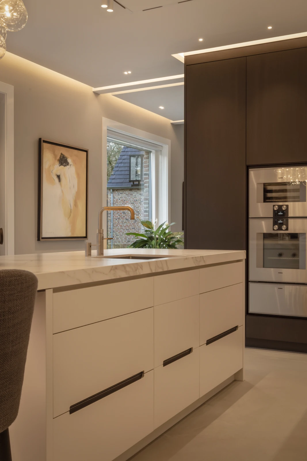 Een moderne keuken ontworpen door een bekwame keukenontwerper heeft witte kasten, ingebouwde roestvrijstalen apparatuur, een marmeren aanrechtblad, een groot raam, hedendaagse kunst aan de muur en een plant in de vensterbank.