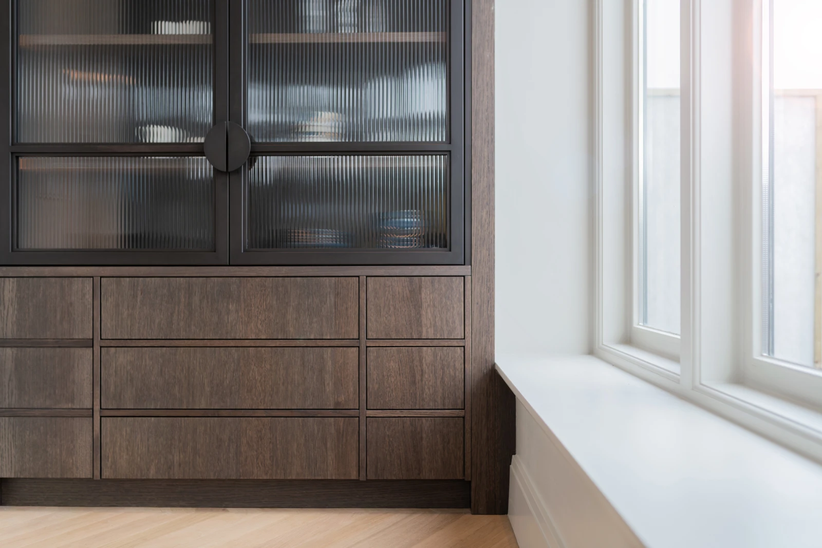 Een moderne houten kast met geribbelde glazen deuren en meerdere laden, geïnspireerd op maatwerk keukens en interieur, staat naast een groot wit omlijst raam met een brede vensterbank, waardoor de lichtgekleurde houten vloer van de kamer wordt gevuld met natuurlijk licht.