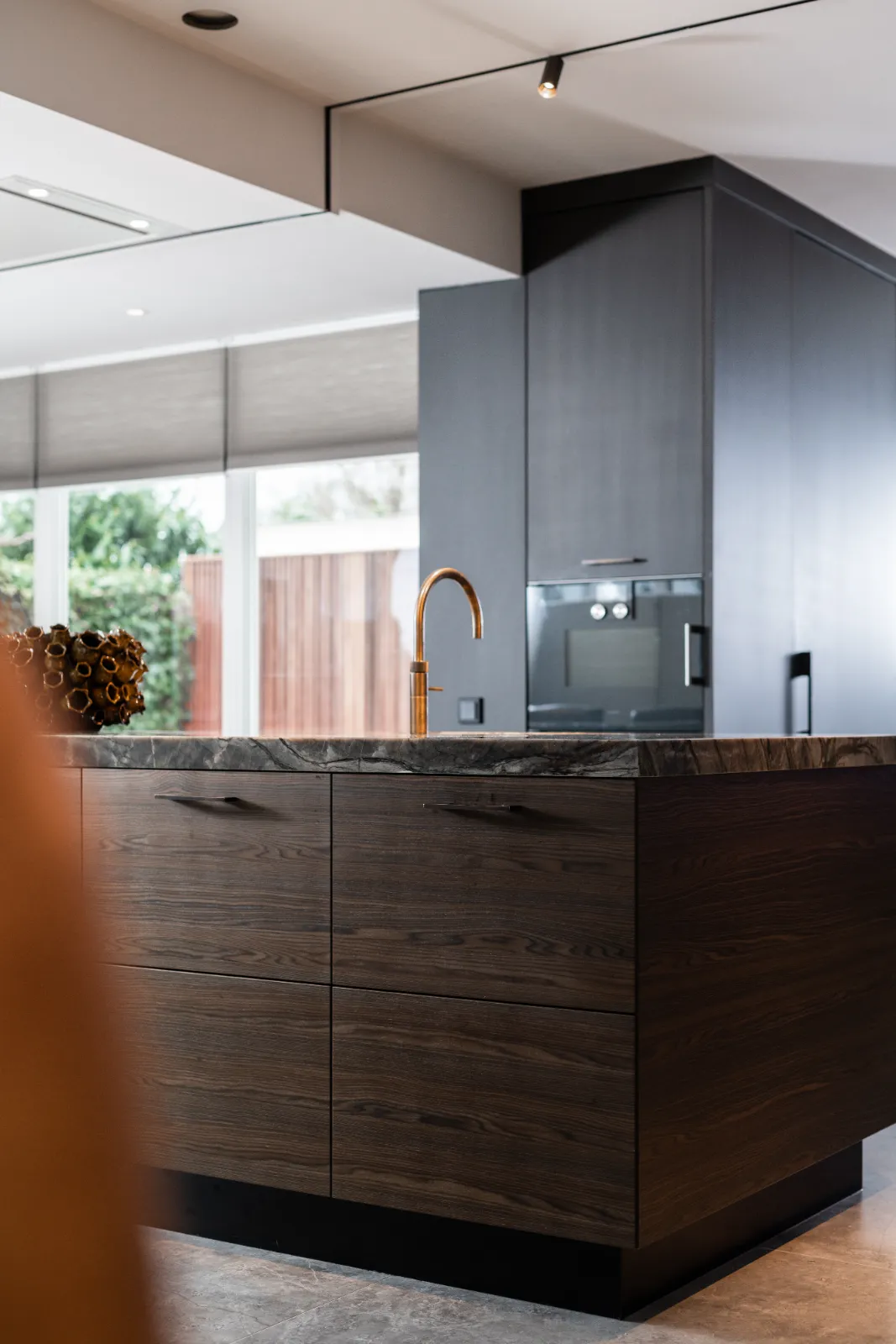 Moderne keuken ontworpen door een gerenommeerde keukenarchitect, met donkerhouten kasten, een marmeren aanrecht en een gouden kraan. Grote ramen op de achtergrond vullen de ruimte met natuurlijk licht; een decoratief voorwerp staat op het aanrecht.