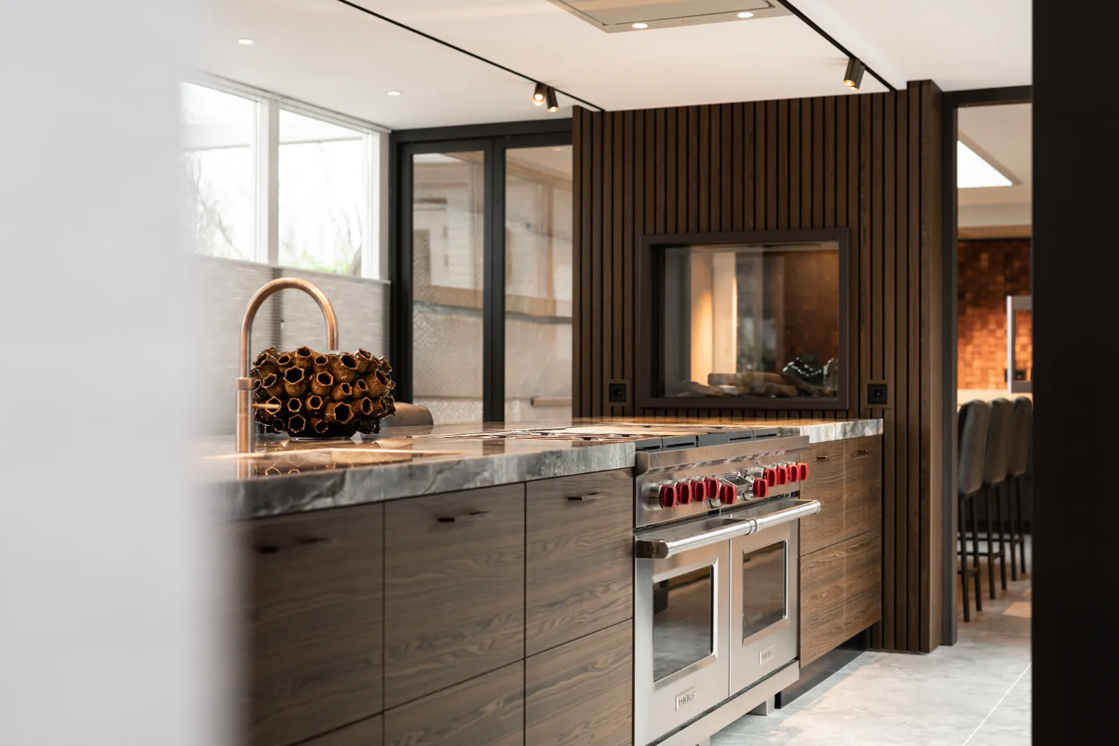 Moderne keuken met houten kasten, marmeren werkbladen en een dubbele oven met rode knoppen, ontworpen door een bekwame keukenarchitect. Een decoratief middenstuk en inbouwverlichting maken de strakke, eigentijdse sfeer compleet.