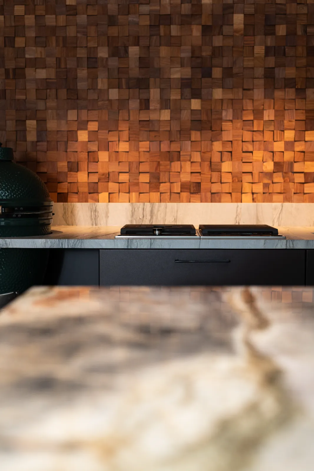 Moderne keuken ontworpen door een keukenarchitect, met een gestructureerde houten blokachterwand, marmeren werkblad, donkere kasten en een groene keramische grill aan de linkerkant. Warme verlichting creëert een gezellige sfeer.