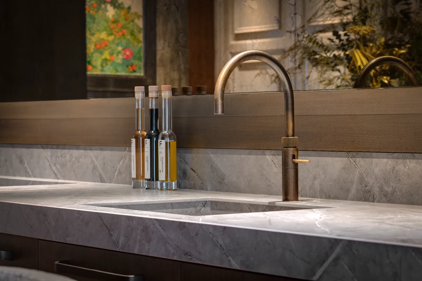 Een moderne wastafel met een geborsteld metalen kraan, een marmeren aanrecht en drie heldere flessen met houten doppen bij de spiegel. Groen en een schilderij worden weerspiegeld in de spiegel, wat keuken herenhuis charme toevoegt aan de ruimte.