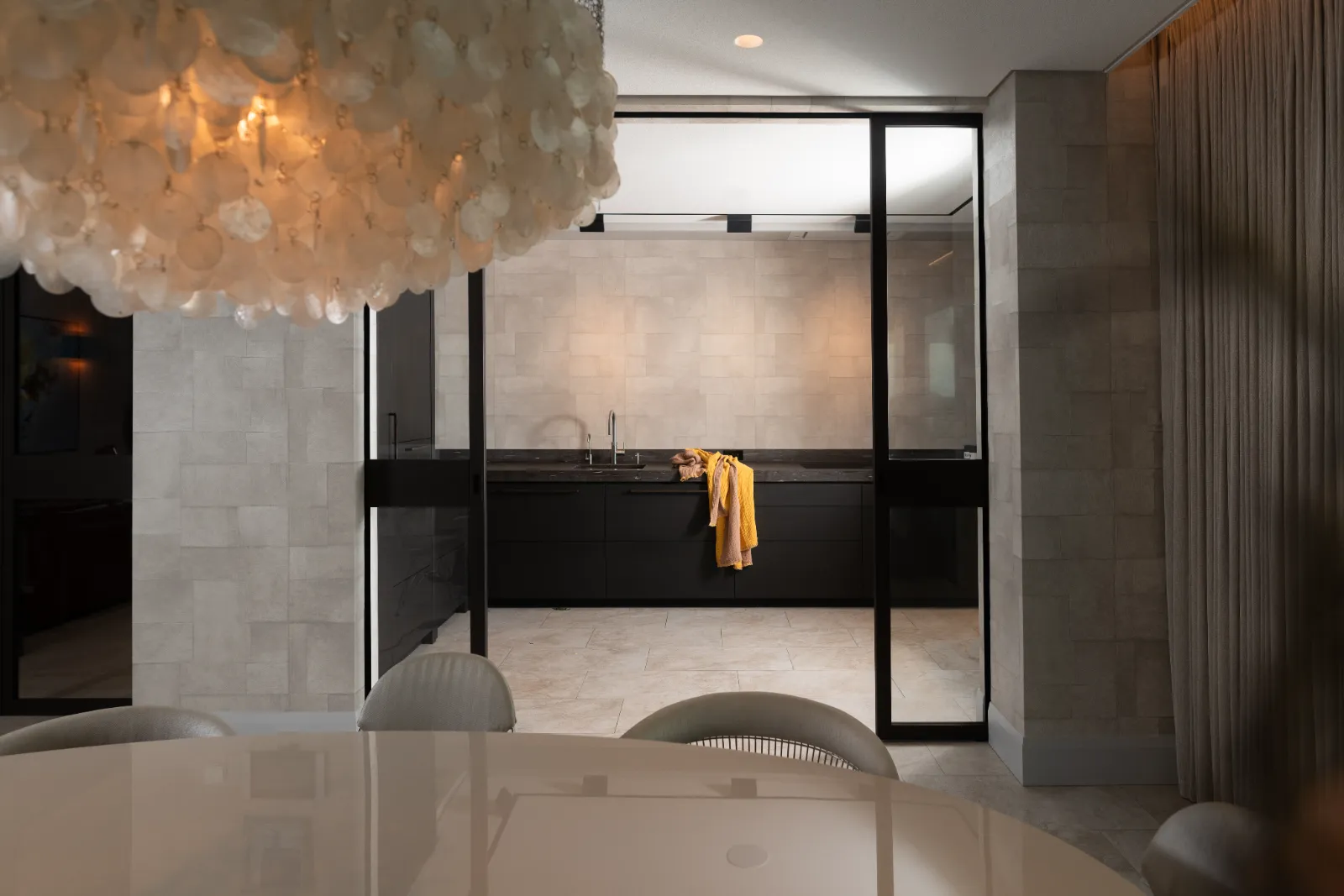 Een moderne keuken met glazen wanden en zwart badmeubel, met een gele handdoek gedrapeerd over het aanrecht. Op de voorgrond, bij het keuken herenhuis, staan een ronde witte tafel en stoelen onder een gestructureerde lamp.