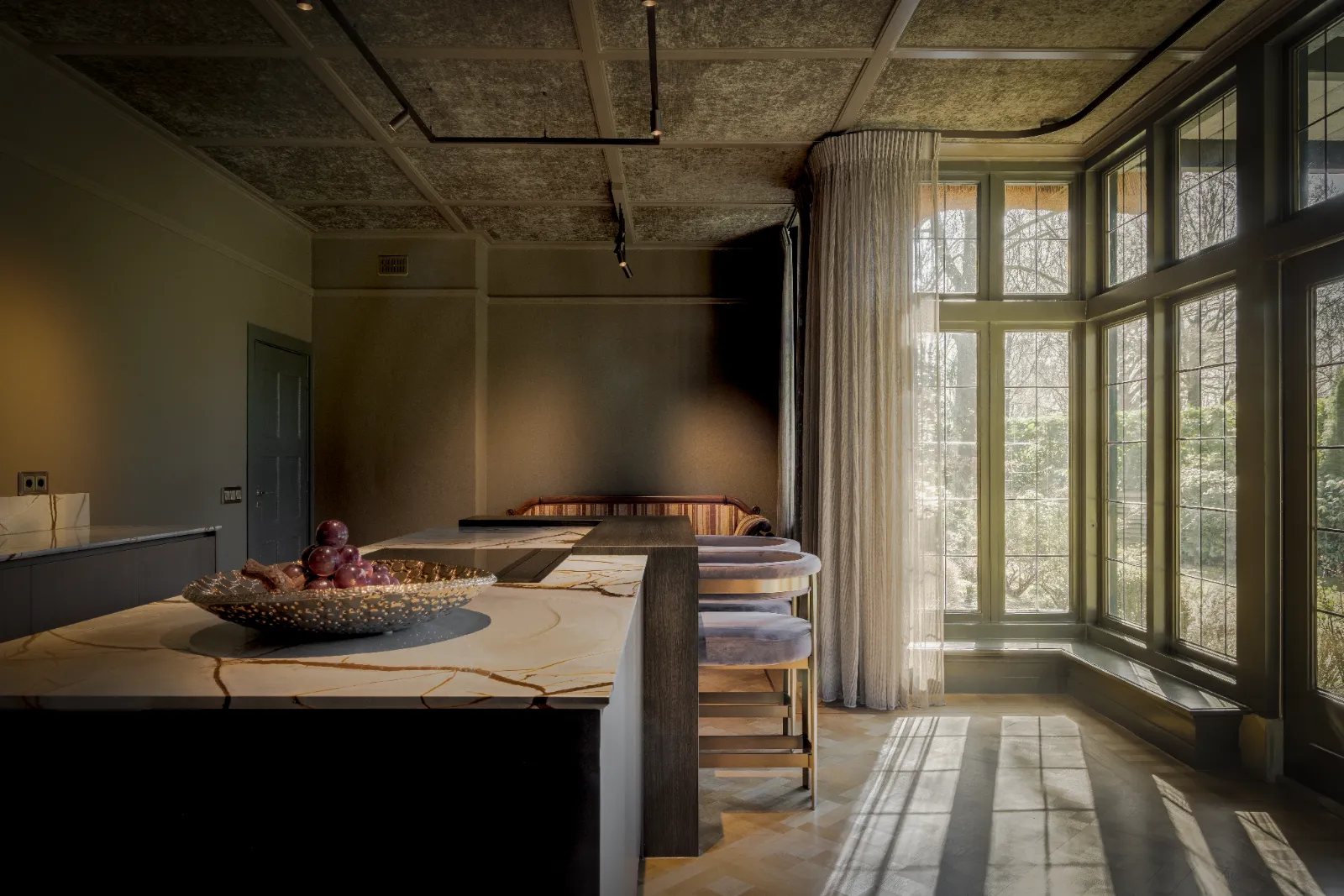 Een modern keuken herenhuis heeft een ruime keuken en eethoek met een groot raam, een marmeren eiland versierd met een decoratieve schaal, pluche barkrukken en zachte gordijnen die het uitzicht naar buiten prachtig omlijsten.