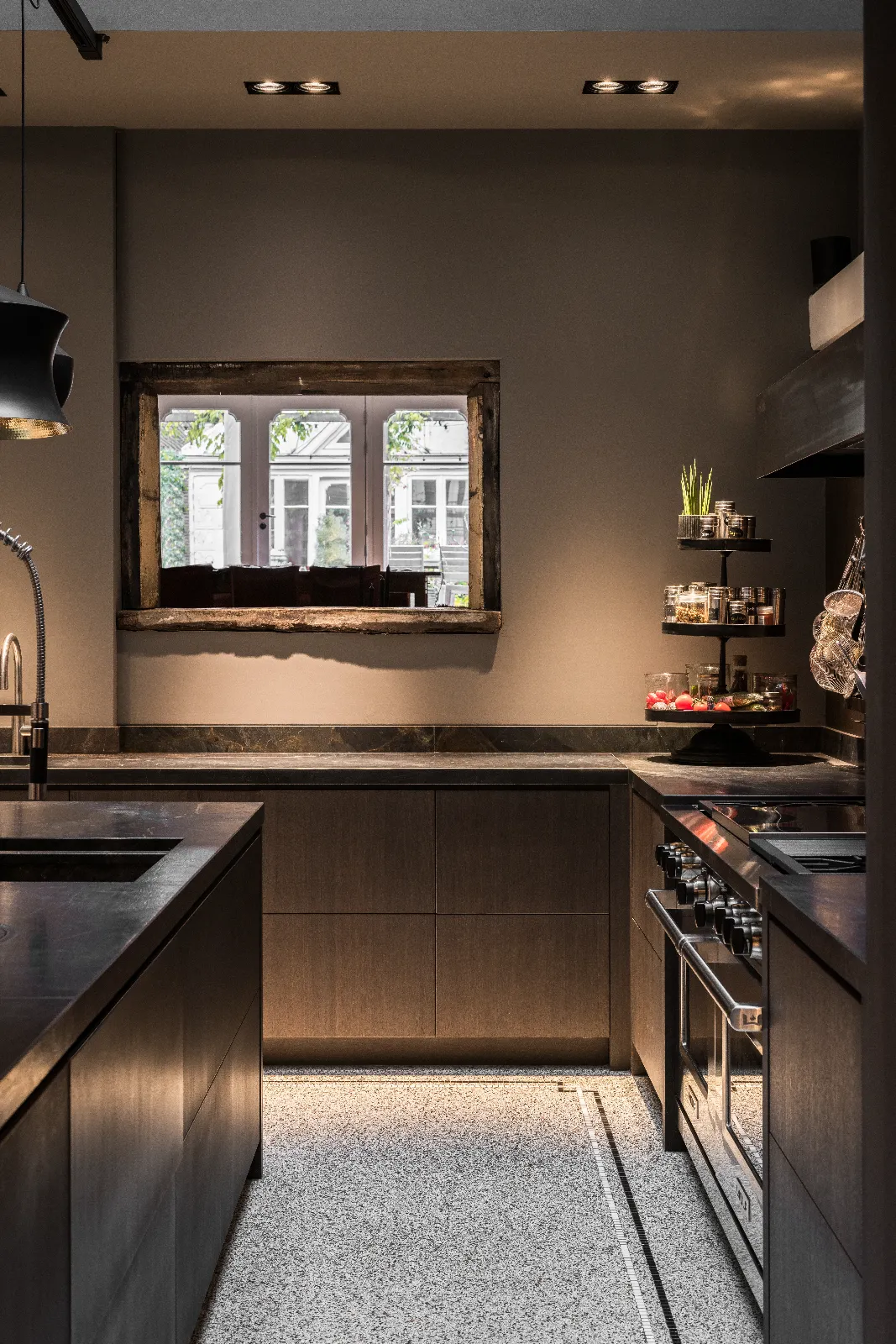 Modern keuken herenhuis met donkere houten kasten, een raam omlijst met rustiek hout, een ingebouwde oven, schappen op niveaus voor planten en kruiden en verlichting onder de kasten die de gespikkelde vloer verlicht.