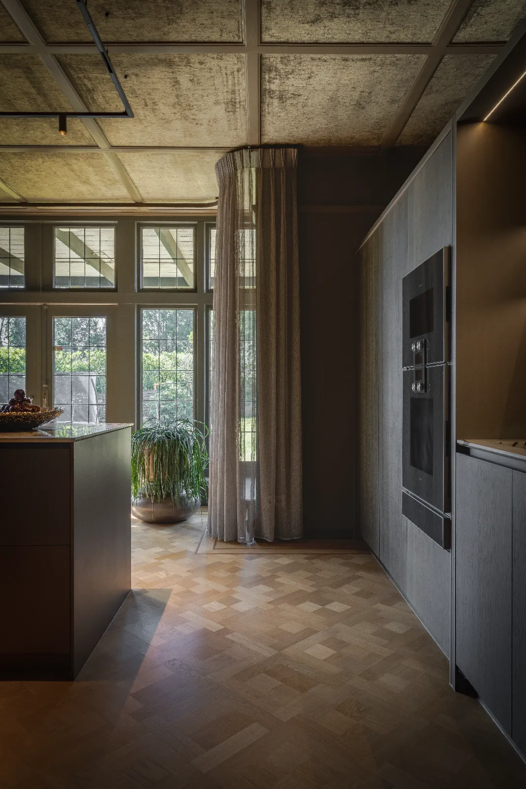 Een modern keuken herenhuis met grote ramen, transparante gordijnen, houten vloeren en inbouwapparatuur; zonlicht valt naar binnen en verlicht een potplant bij het raam.