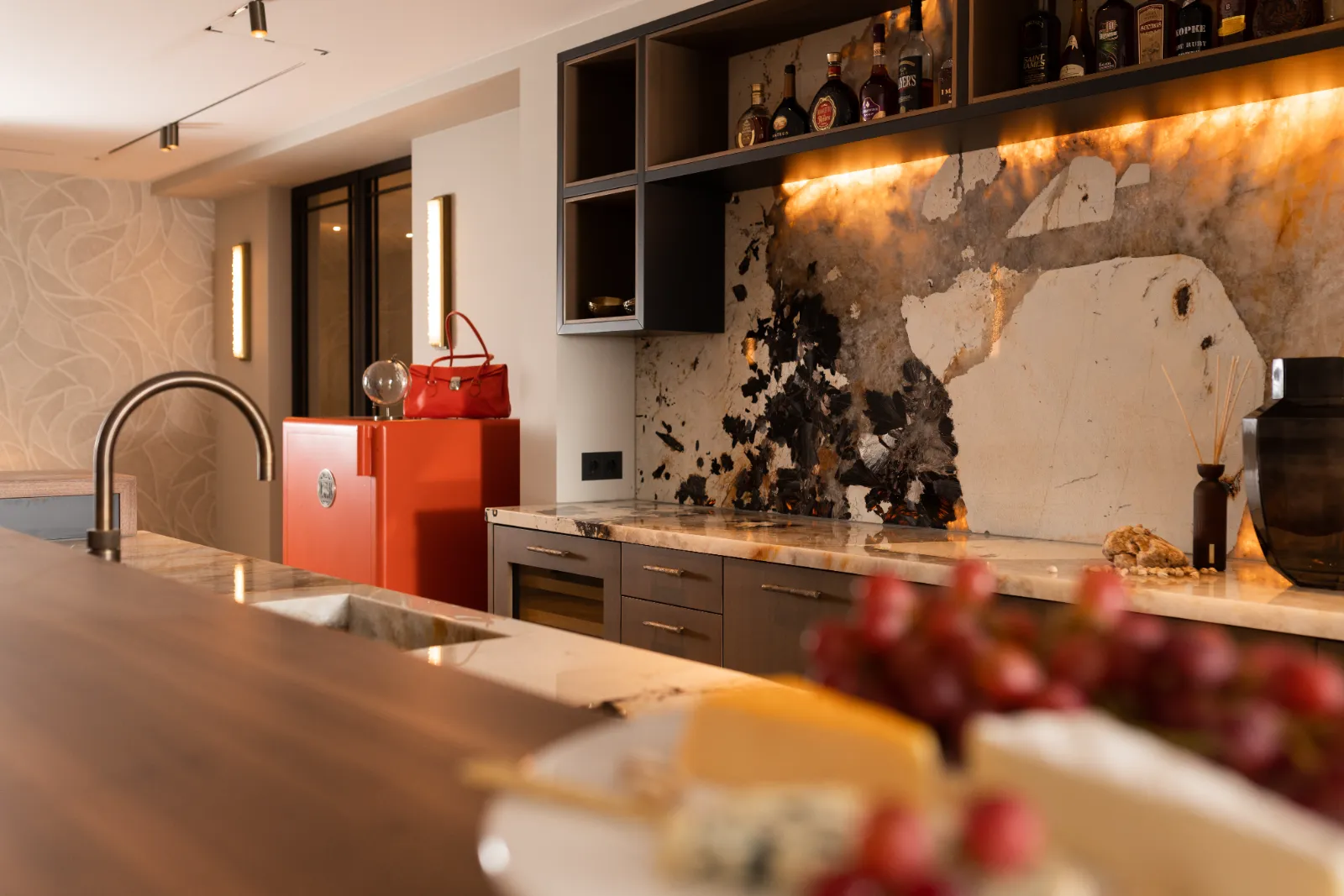 Een moderne keuken ontworpen door een bekwame keukenontwerper heeft een marmeren achterwand, houten kasten, verlichting onder de schappen en uitgestalde flessen. Een rode koelkast met een bijpassende handtas staat bij het aanrecht, terwijl een kaasplankje met druiven ervoor staat.