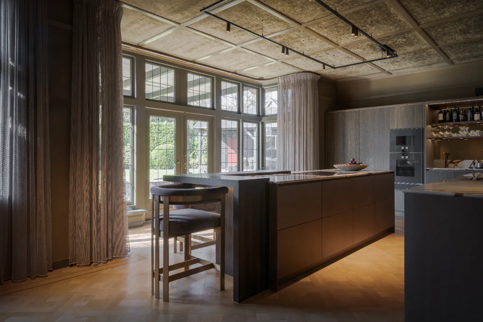 Moderne keuken met grote ramen, ontworpen door een ervaren keukenontwerper, die natuurlijk licht binnenlaten. Centraal eiland met stoelen, een fruitschaal en strakke apparatuur. Lichte houten vloeren, hoge gordijnen en groen buiten zorgen voor een warme, uitnodigende sfeer.