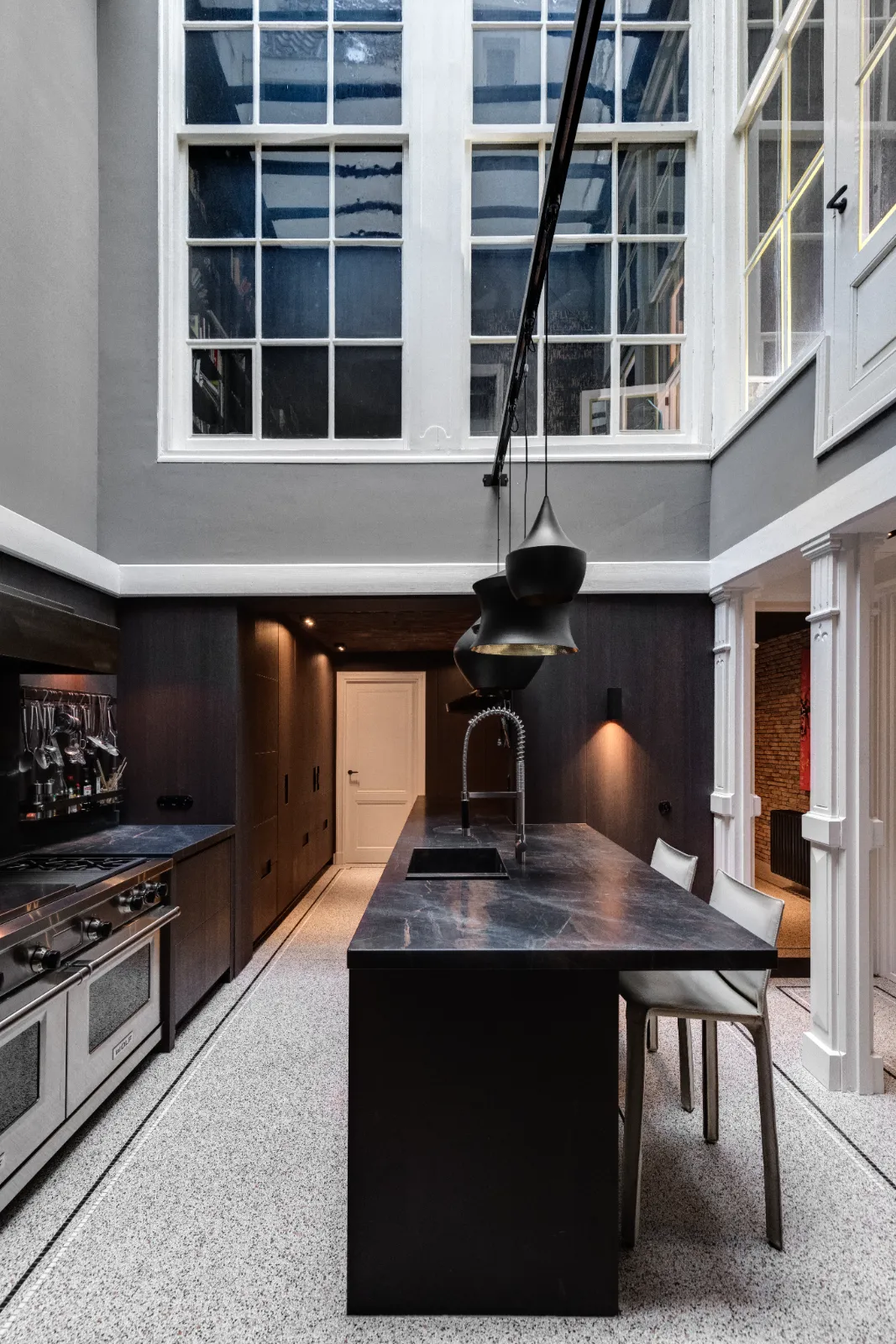 Moderne keuken ontworpen door een ervaren keukenontwerper, met een zwart marmeren eiland, hoge plafonds, grote ramen, minimalistische krukken, hangende hanglampen, ingebouwde apparatuur, donkere kasten die contrasteren met witte muren en terrazzovloeren.