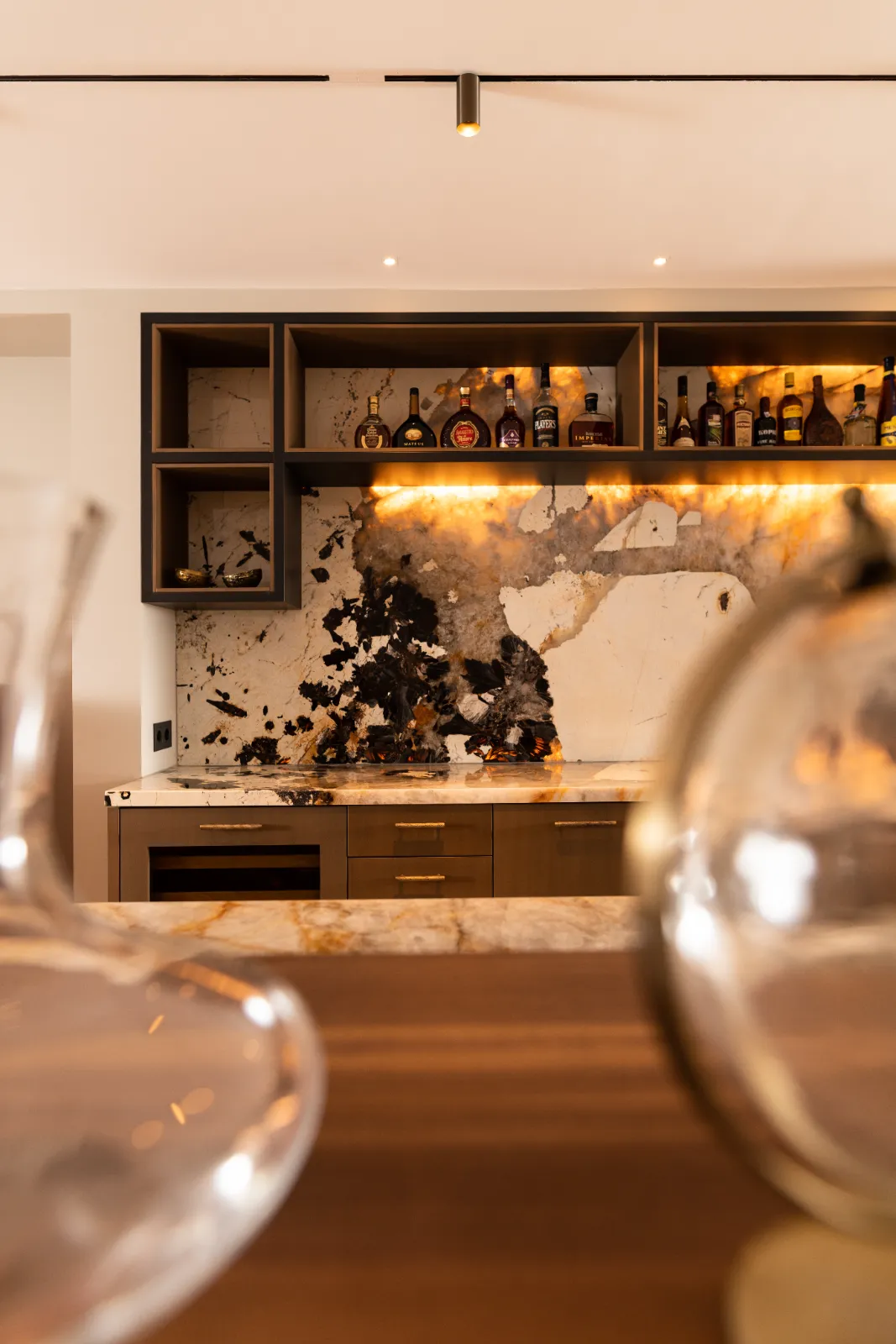 Moderne huisbar met een marmeren achterwand, ontworpen door een ervaren keukenontwerper, met open schappen voor drankflessen, warme verlichting onder de kast en een houten aanrechtblad. Decoratieve glazen voorwerpen zijn vaag op de voorgrond.