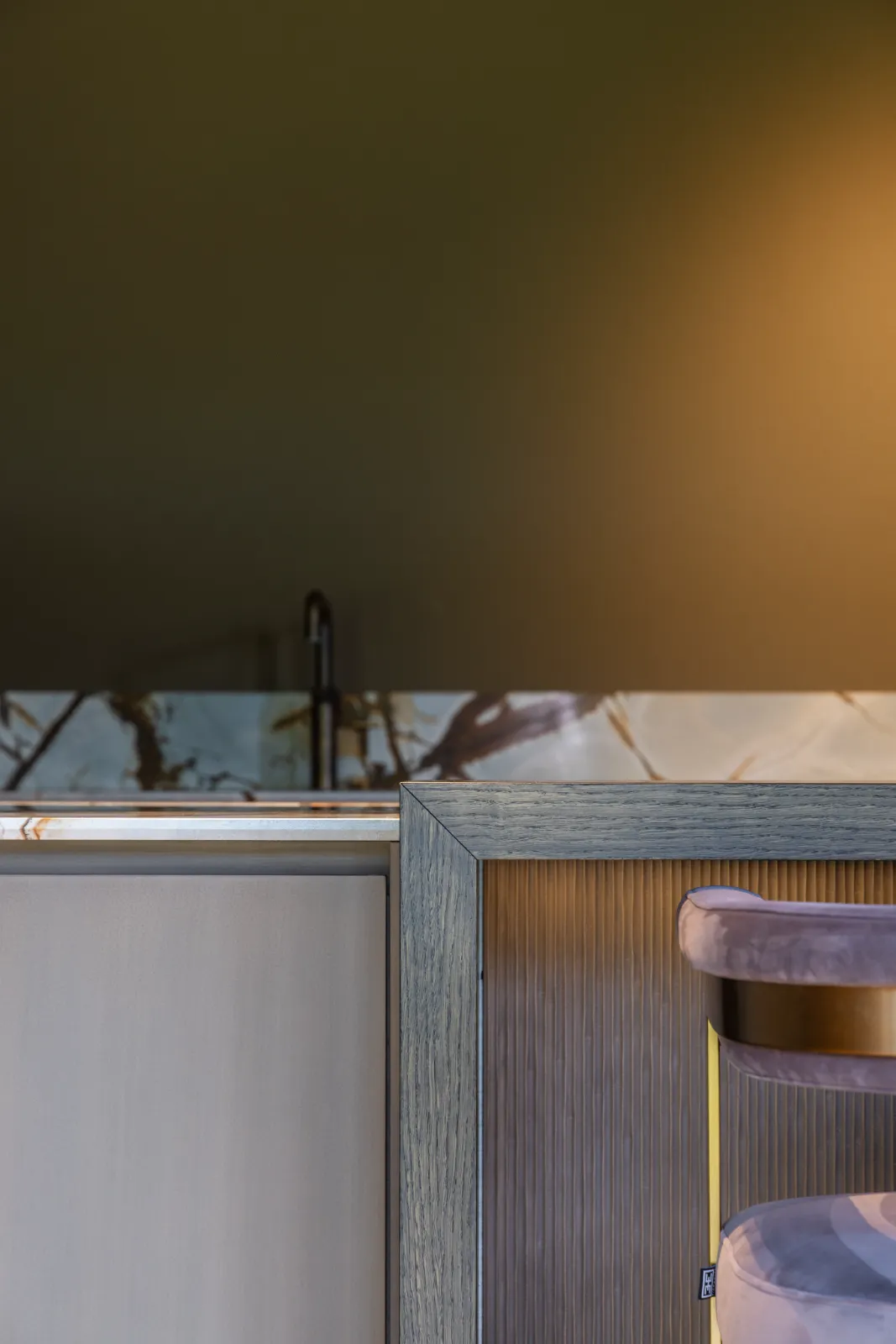 Een moderne keuken van een bekwame keukenontwerper heeft een marmeren achterwand, een minimalistische kraan, een houten kast en een deel van een met fluweel beklede stoel met een gouden frame op de voorgrond.