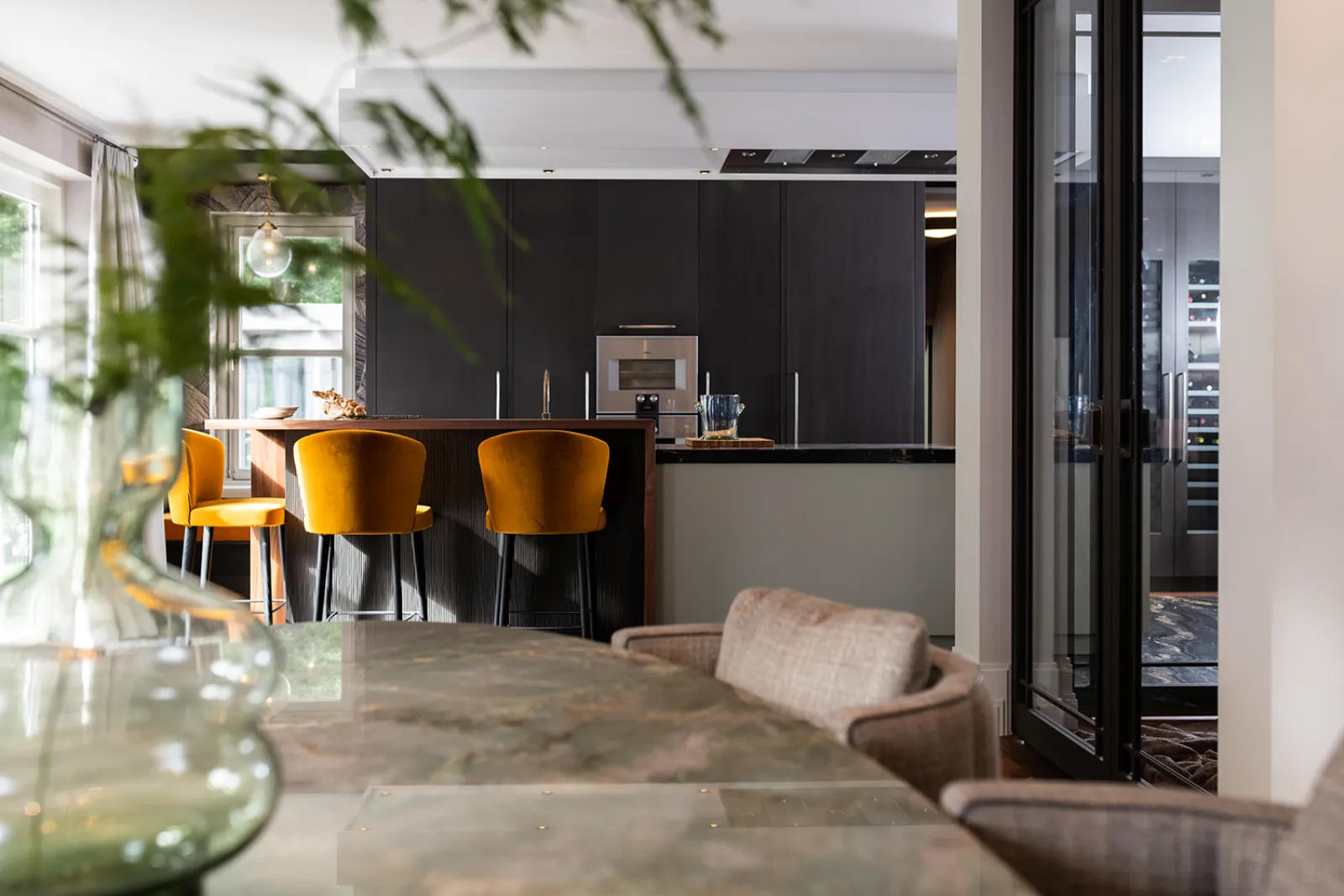 Moderne keukenvilla met een marmeren eettafel, beige gestoffeerde stoelen en gele barkrukken aan een donker keukeneiland. Een glazen vaas met groen gebladerte staat op de voorgrond terwijl het natuurlijke licht door de grote ramen naar binnen valt.