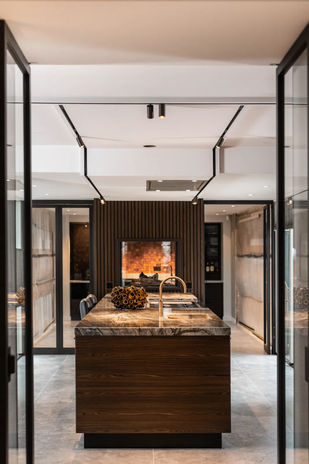 Een moderne keuken in een keukenvilla met een centraal eiland met een marmeren aanrechtblad, ingebouwde gootsteen en vaas met gedroogde bloemen. Op de achtergrond worden een wand met houten panelen en een bakstenen oven zacht verlicht door warme verlichting.