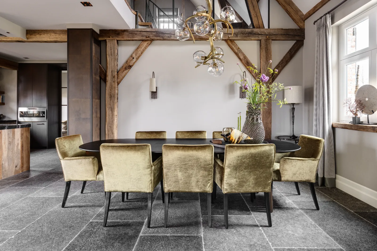 Moderne eetkamer met een donkere ovale tafel, acht goudfluwelen stoelen, een sculpturale kroonluchter, zichtbare houten balken, stenen vloertegels, groot raam met gordijnen en decoratieve vazen met bloemen op de tafel en bijzettafel.