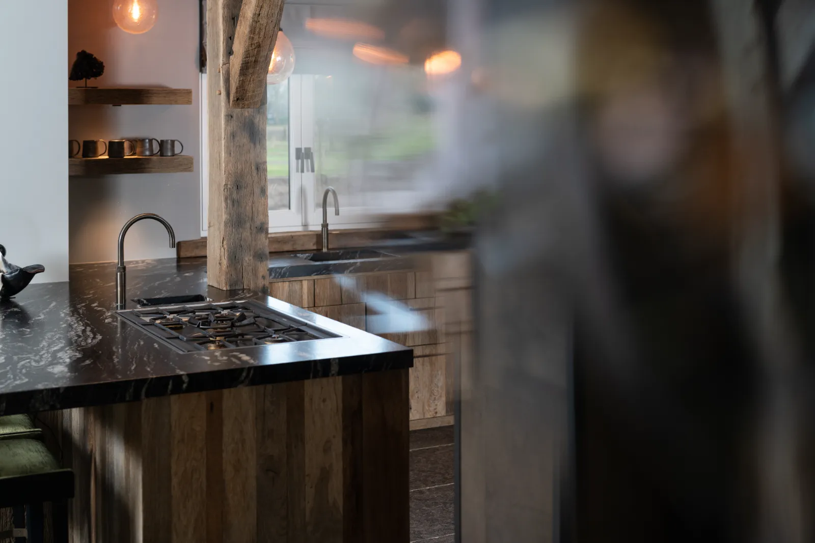 Een moderne, rustieke keuken met houten kastjes, een zwart marmeren aanrechtblad, een ingebouwd gasfornuis, een kleine kraan, open schappen met kopjes en grote ramen die natuurlijk licht binnenlaten.