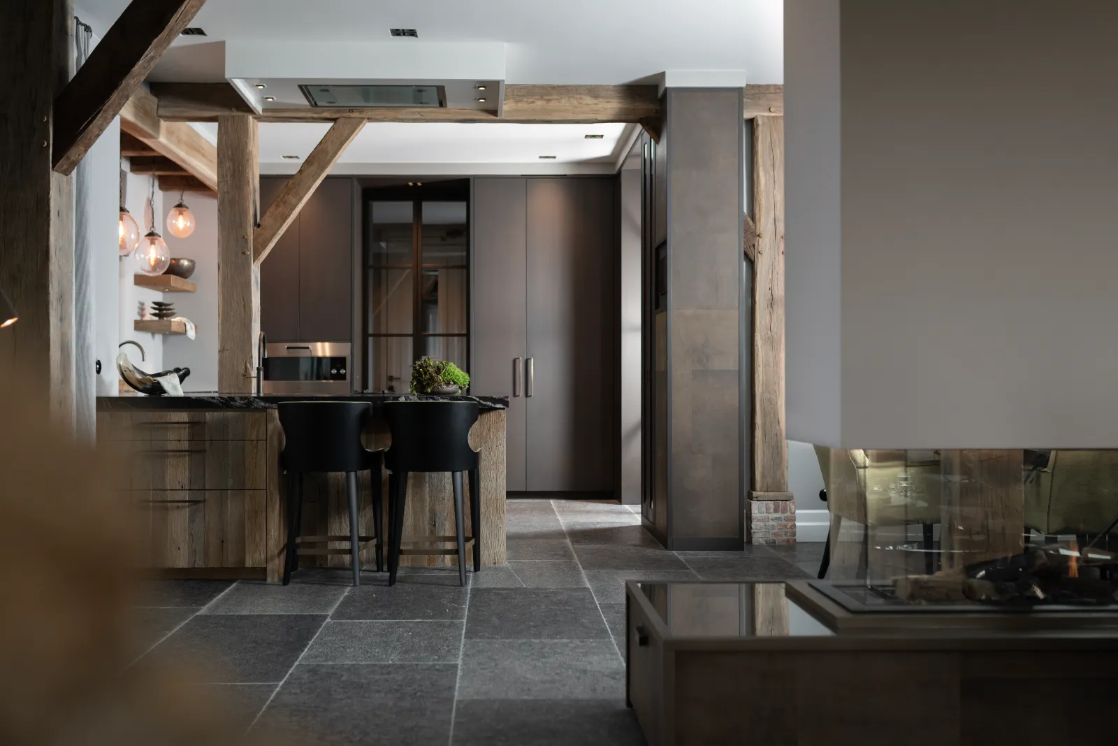Moderne rustieke keuken woonboerderij met donkere kasten, houten balken, een zwart eiland met twee stoelen, stenen tegelvloer en hedendaagse verlichting. Een met glas afgesloten open haard en eethoek zijn gedeeltelijk zichtbaar op de voorgrond.