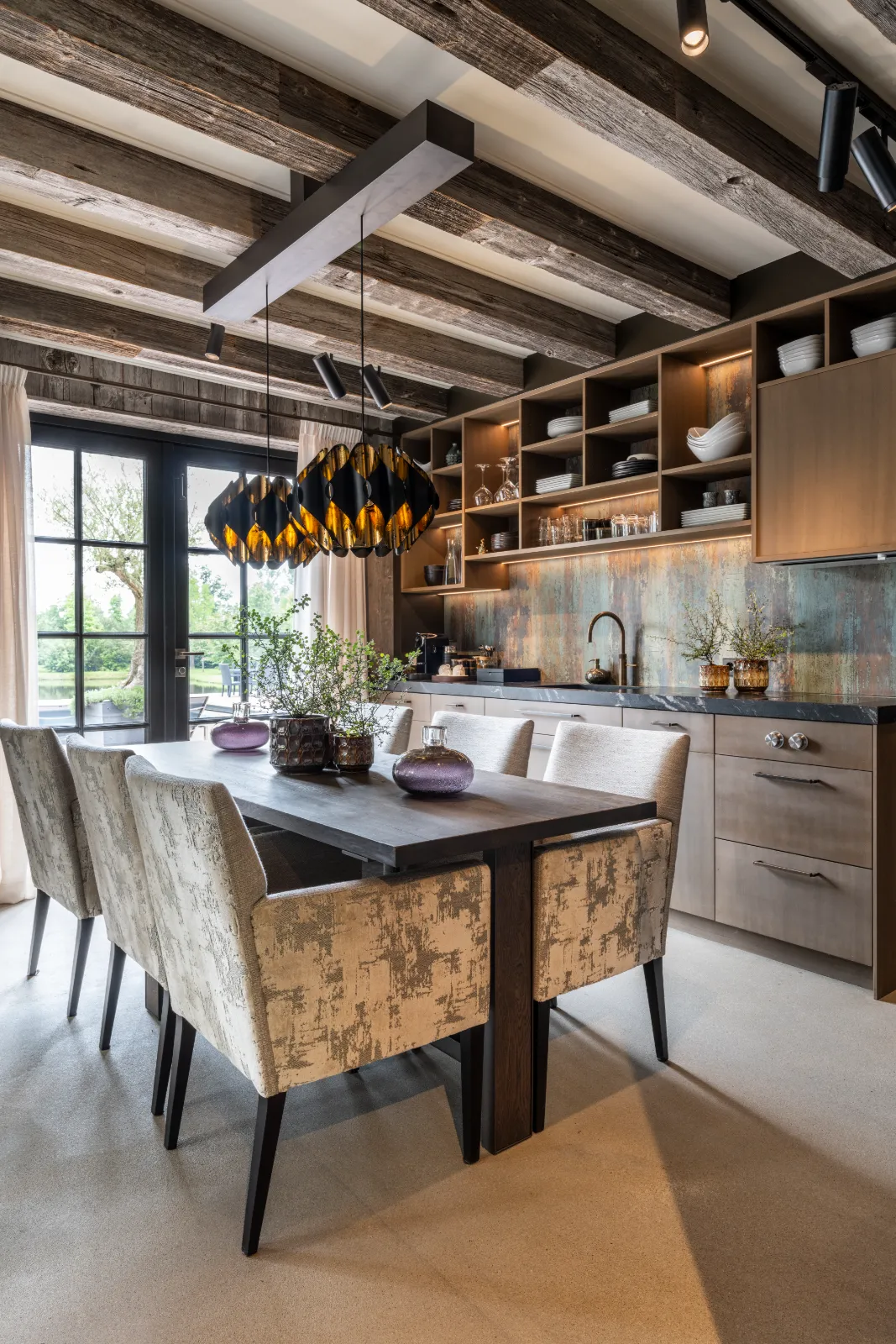 Moderne keuken en eethoek met houten balken vangen de charme van een interieur woonboerderij. Open rekken, een rustieke houten tafel en grote hanglampen creëren een gezellige ruimte met glazen deuren die weelderig groen buiten omlijsten.