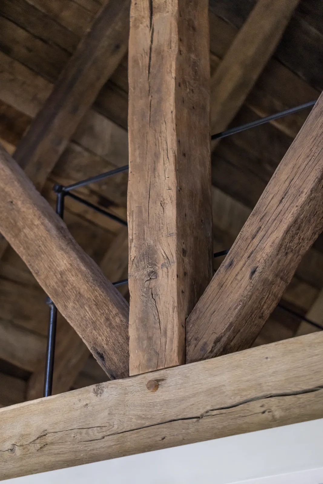 Close-up van rustieke houten balken met zichtbare nerven en knoesten, samengevoegd in een plafondconstructie van een woonboerderij. Dunne zwarte metalen staven kruisen elkaar achter het hout en geven steun aan het bijpassende houten plafond erboven.