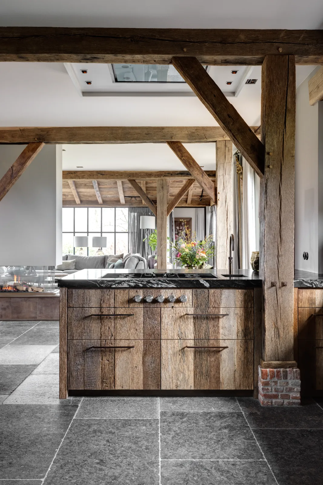 Een rustieke keuken, geïnspireerd op woonboerderij interieur, heeft verweerde houten kasten, zichtbare houten balken, een stenen tegelvloer en een donker aanrechtblad. De ruimte loopt over in een lichte woonkamer met grote ramen en veel groen.