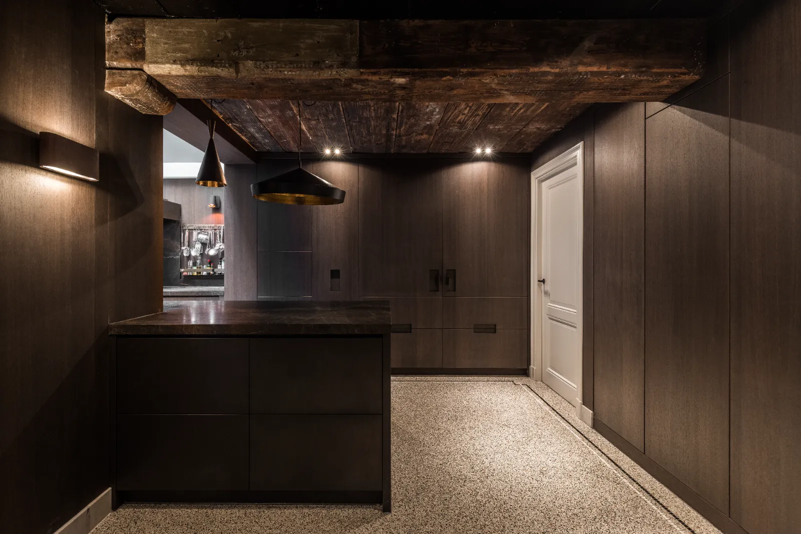 Minimalistische keuken met donkerhouten kasten, een zwart eiland en moderne hanglampen. Zichtbare rustieke balken benadrukken het maatwerk interieur en keuken, terwijl een witte deur en gespikkelde vloer contrasteren met het strakke, stemmige interieur.