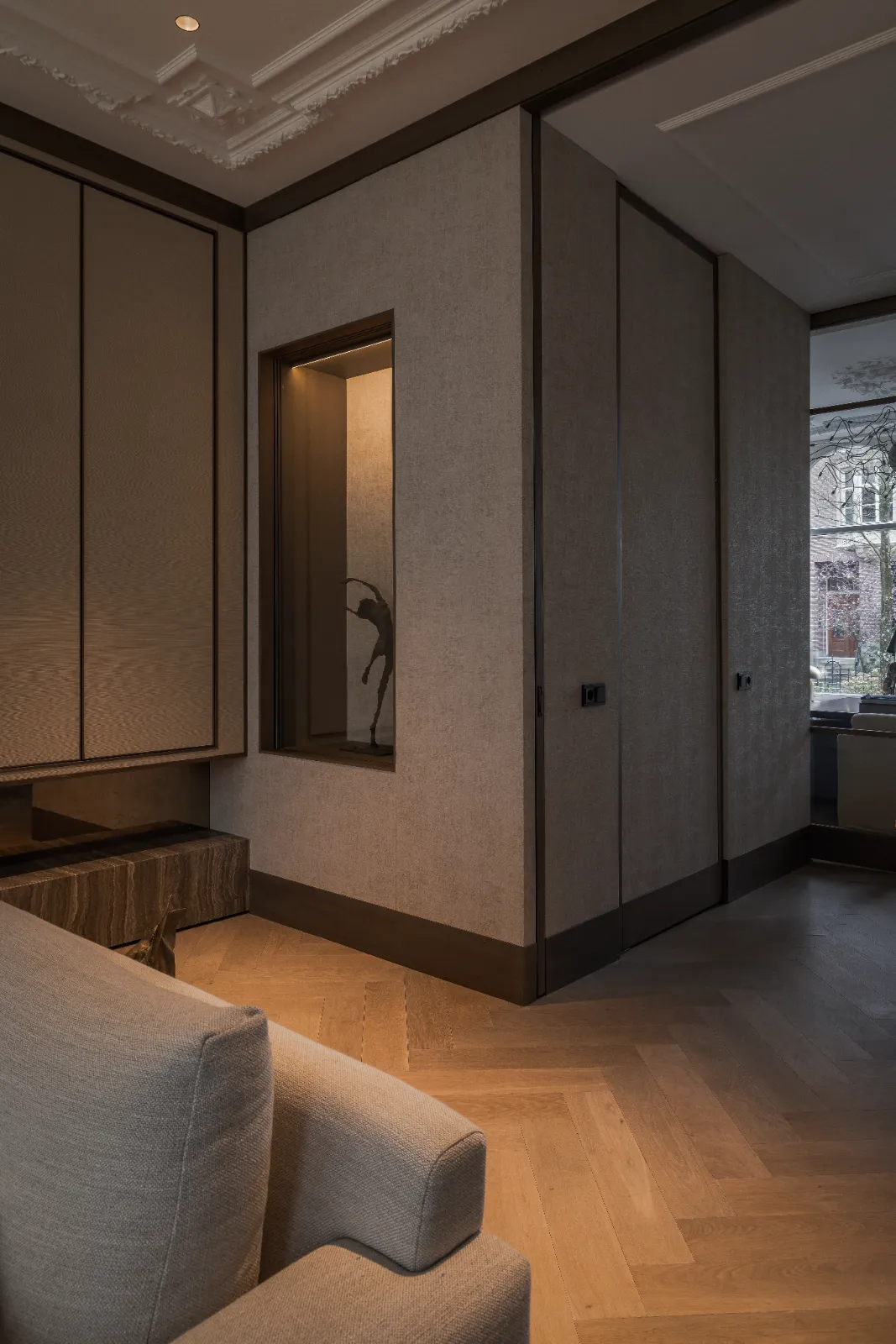 Een moderne, gedimd verlichte kamer met gedempte grijze muren, houten visgraatvloer en een lichtgrijze bank laat maatwerk interieur en keuken zien. Een grote ingebouwde vitrine met een donker beeldhouwwerk en een raam met uitzicht op de bomen buiten maken het plaatje compleet.