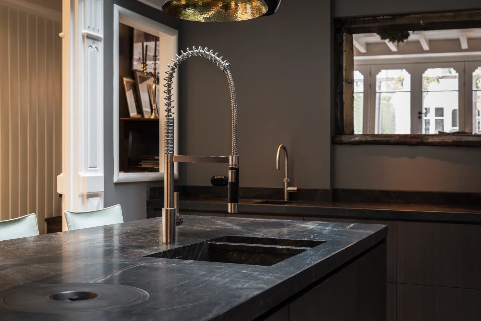 Moderne maatwerk keuken met een donker marmeren aanrechtblad, een grote industriële veeruittrekkraan en een vierkante spoelbak. Op de achtergrond maken een tweede kleinere kraan en een raam dat licht weerkaatst het ontwerp compleet.