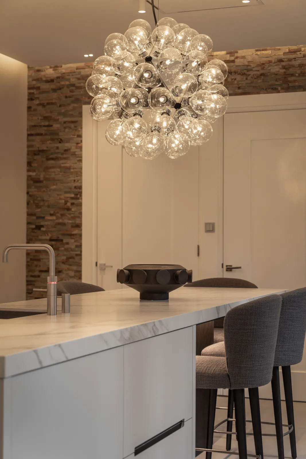 Moderne maatwerk keuken met een marmeren eiland, grijs gestoffeerde barkrukken, een strakke kraan en een cluster bolvormige hanglampen. De achtergrond is voorzien van een accentmuur van structuursteen en witte dubbele deuren.