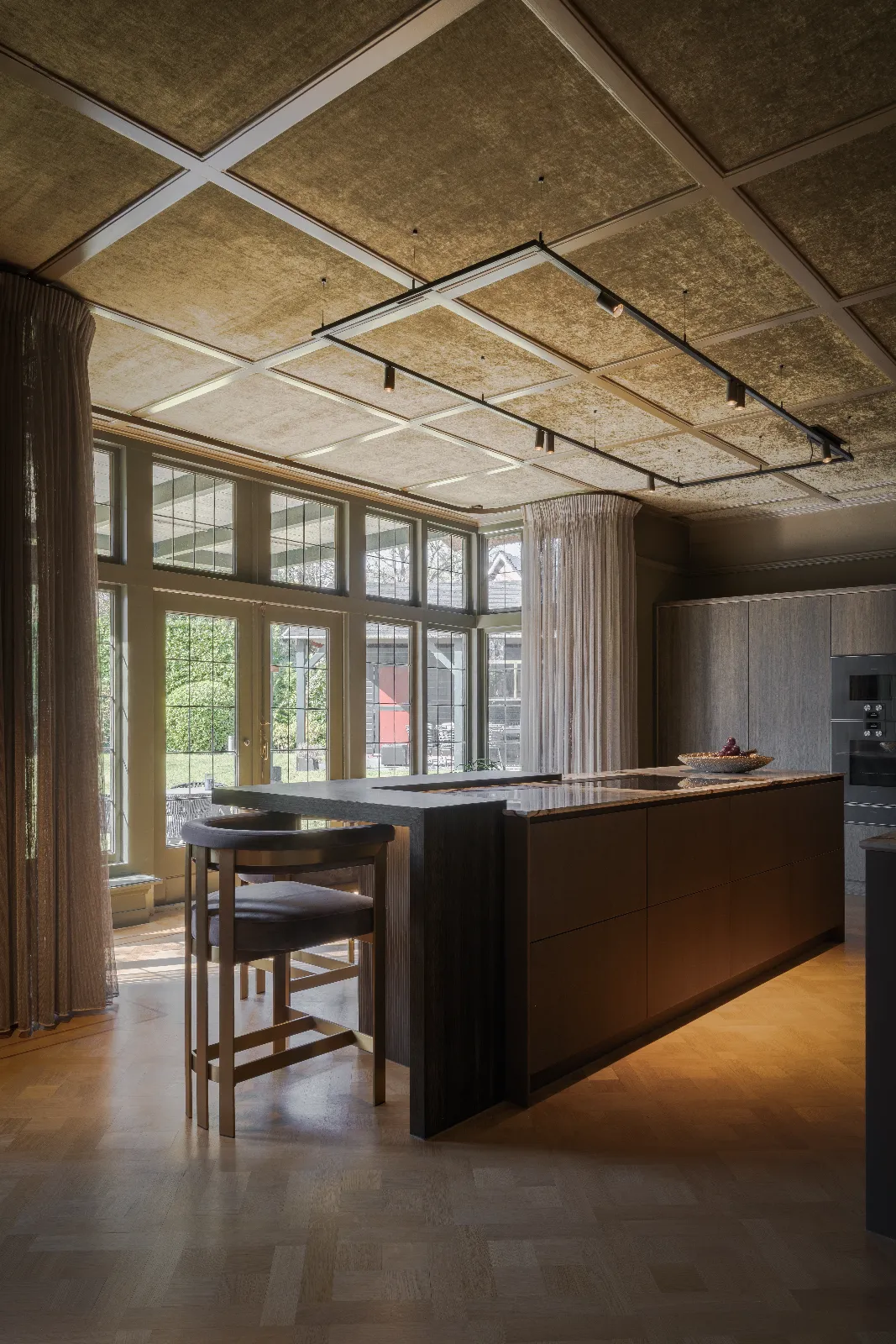 Moderne maatwerk keuken met grote ramen die daglicht binnenlaten, een centraal donkerhouten eiland met barkrukken, parketvloeren en transparante gordijnen. Buiten zijn bomen en een rode structuur zichtbaar.