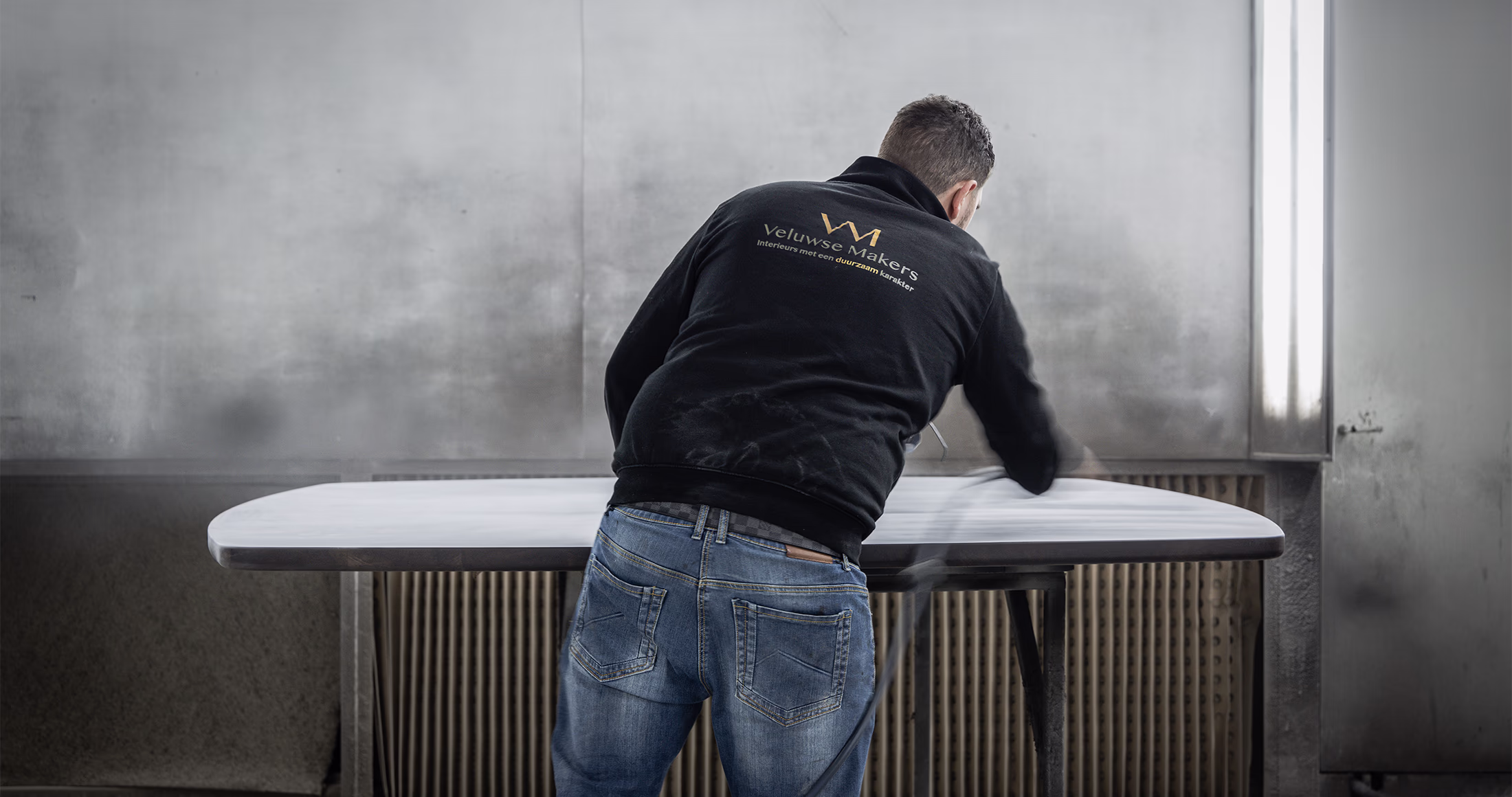 Een persoon met een zwarte Veltrup Mobel jas is een grote, ovale tafel aan het polijsten of schoonmaken in een werkplaats of industriële omgeving. De persoon kijkt weg van de camera.